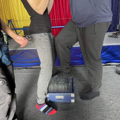 Schüler steigt auf das Nagelbrett. Lehrerin unterstützt sie. 