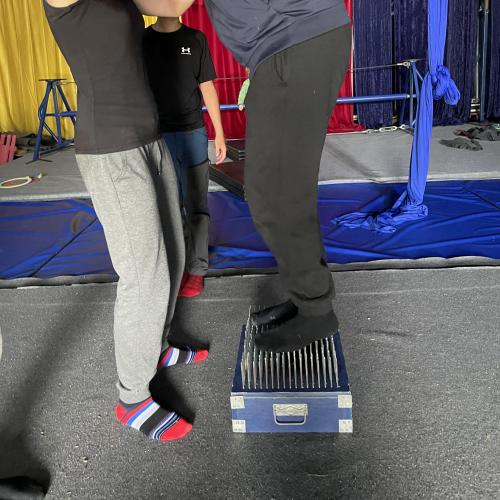 Schüler steigt auf das Nagelbrett. Lehrerin unterstützt sie. 